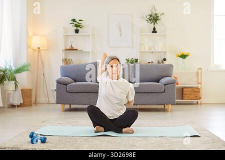 Donna matura flessibile seduta sul tappetino da yoga a casa e che fa esercizi di stretching Foto Stock