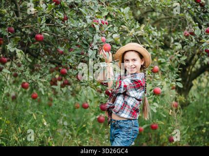 Una ragazza allegra si erge sullo sfondo di un meleto con mele rosse, gusta spuntini, vitamine e frutta biologica. Lei è vestita in scacchi shir Foto Stock