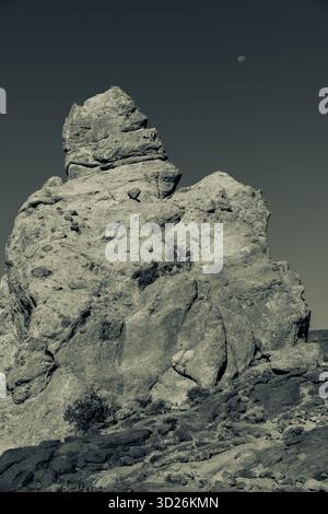 Immagine in bianco e nero di un alto pinnacolo di roccia frastagliato che si innalza contro un cielo scuro con una luna visibile sopra e arbusti sparsi sulla base rocciosa Foto Stock