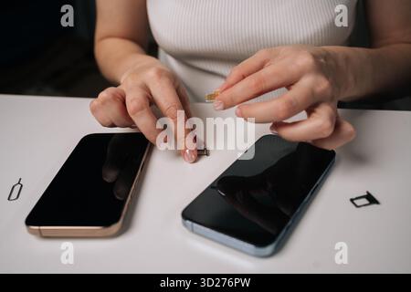 Vista dall'alto della donna che inserisce la scheda sim nel nuovo smartphone, con un altro telefono e uno strumento di espulsione sim nelle vicinanze su un tavolo bianco, durante la configurazione del dispositivo mobile Foto Stock