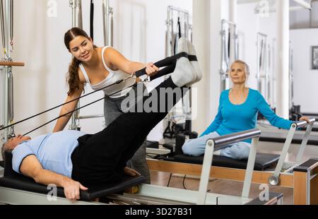 Giovane istruttrice positiva che controlla i movimenti dell'uomo anziano che si allunga sulle attrezzature del riformatore Pilates in studio Foto Stock