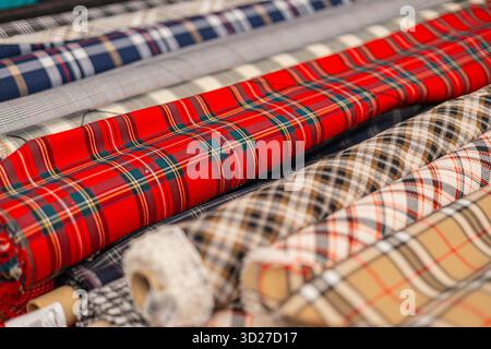 Primo piano di rotoli di tessuto plaid impilati con motivi tartan, caratterizzati da audaci scacchi rossi, beige, grigi e multicolori. Le texture variano da nitide a morbide Foto Stock
