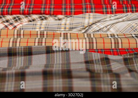 Primo piano di rotoli di tessuto plaid impilati con motivi tartan, caratterizzati da audaci scacchi rossi, beige, grigi e multicolori. Le texture variano da nitide a morbide Foto Stock