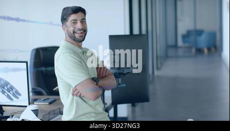 Uomo indiano in piedi in una polo verde chiaro che incrocia le armi in ufficio tecnologico, monitor, spazio fotocopie Foto Stock