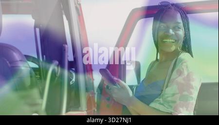 Donna nera sorridente che tiene lo smartphone davanti alla porta rossa del SUV parcheggiata, con volante e cruscotto Foto Stock