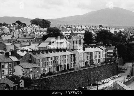 Annidata sotto le imponenti mura del Castello di Caernarfon, la città di Caernarfon rivela un'affascinante miscela di architettura storica e vita quotidiana. File di case a schiera, prevalentemente di color crema e giallo pallido, scendono lungo la collina, offrendo un pittoresco scorcio sulla routine quotidiana gallese. Il paesaggio circostante, dominato dalla lontana catena montuosa Snowdonia, completa questa iconica scena, mostrando la posizione strategica della città e la sua bellezza duratura. Foto Stock