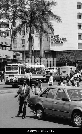 Le vivaci strade di Nairobi si animano di vita. Questa immagine in bianco e nero cattura un momento nel cuore della città, mostrando lo Stanley Hotel e il traffico vivace lungo la strada. Un uomo in tuta si ferma in mezzo ai veicoli, una testimonianza della miscela di affari e vita quotidiana della città. La scena riflette l'energia dinamica di Nairobi e il ricco arazzo culturale. Foto Stock