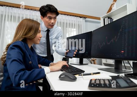 Uomo d'affari asiatico sorridente che mostra dati dinamici del mercato azionario al telefono con una donna che cerca al computer per analizzare il tasso di cambio del valore del profitto o Foto Stock