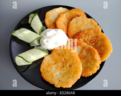 Frittelle di patate servite con panna acida e fette di cetriolo su un piatto nero. Foto Stock