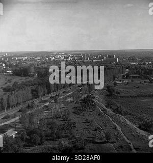 Archivio foto in bianco e nero scattata a Sloviansk, Ucraina, nell'agosto 1997. Si tratta di una vista panoramica ad alto angolo della citta', presa dalla SKTB (Special Design and Technological Bureau). Lo scatto cattura un vasto paesaggio urbano, mostrando i quartieri residenziali della città all'orizzonte, compresi gli edifici a più piani. In primo piano, è visibile una grande zona verde, possibilmente un parco o una cintura forestale, con una strada principale che corre accanto ad essa. Questa immagine documenta l'aspetto della città alla fine degli anni '1990 Foto Stock