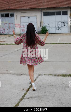 Vista posteriore di una donna con un abito floreale e tacchi alti bianchi che camminano all'aperto su una superficie di cemento vicino a un vecchio edificio con graffiti alle pareti. Foto Stock