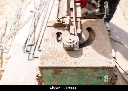 Un lavoratore che indossa guanti rossi aziona l'attrezzatura in metallo arrugginito per piegare le barre di rinforzo in acciaio, con altre barre di armatura e materiali da costruzione Foto Stock