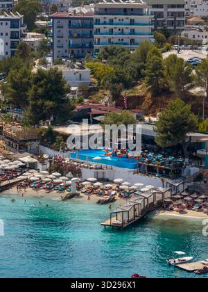 Vista aerea delle acque cristalline turchesi che incontrano la spiaggia sabbiosa, punteggiata di lettini al sole, vicino agli edifici vibranti di Ksamil, Vlor, Albani Foto Stock