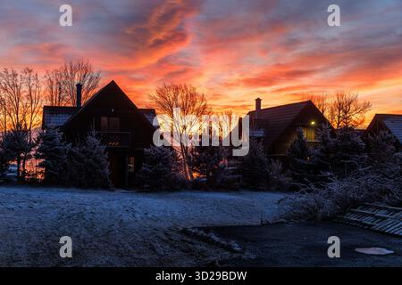 Le case ricoperte di gelo si trovano sotto un cielo luminoso all'alba in un tranquillo insediamento di montagna. Le nuvole calde contrastano con il terreno ghiacciato. Foto Stock