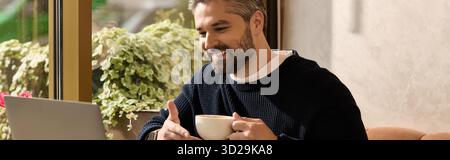 Un bell'uomo con un abbigliamento elegante sorride e sorride mentre lavora al suo computer portatile al bar. Foto Stock