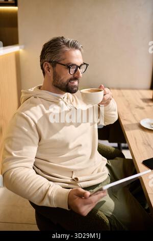 Un uomo bello e maturo sorseggia caffè e legge sul suo tablet in un moderno ambiente da caffè. Foto Stock