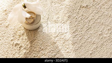 Due fedi nuziali dorate si trovano su un podio rotondo in cemento naturale con un fiore bianco. Sfondo in cemento color sabbia goffrato. spazio per il testo Foto Stock