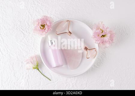 Massaggiatore a rullo al quarzo rosa, raschietto gua Sha, olio per massaggi e trattamenti in bottiglia opaca su piastra in ceramica bianca. Vista dall'alto. Effetto di sollevamento Foto Stock