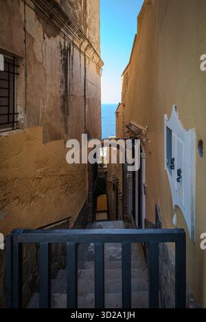 Stretta corsia in discesa con scale tra case sulla scogliera a Positano, Italia, con un frammento di cielo blu e mare visibile in lontananza Foto Stock