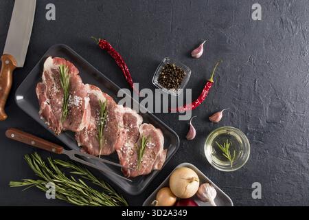 Tre pezzi di collo di maiale con rosmarino, pepe, cipolla, aglio su un vassoio scuro, un coltello e una forchetta su sfondo nero per cucinare. vista dall'alto Foto Stock