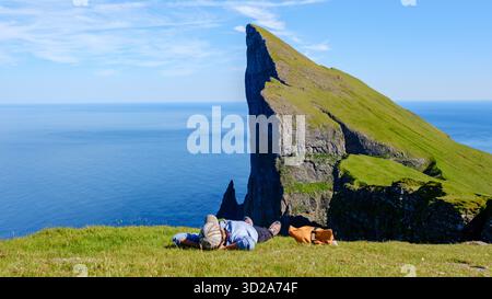 Una donna asiatica si crogiola alla luce del sole su una scogliera erbosa di Mylingur nelle Isole Faroe, godendo di vedute mozzafiato dell'oceano e delle maestose formazioni rocciose che si innalzano bruscamente dal mare. || modello rilasciato Foto Stock