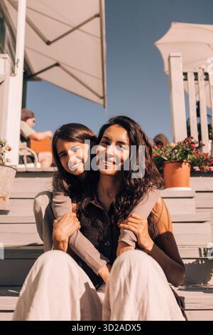 Ritratto di una ragazza sorridente con il braccio intorno alla madre seduta sui gradini Foto Stock