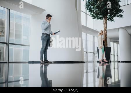 Uomo d'affari al telefono con documenti in una moderna hall mentre un collega aspetta nelle vicinanze Foto Stock
