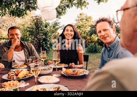 Amici di famiglia sorridenti, uomini e donne, che cenano nel cortile posteriore per una festa di riunione sociale Foto Stock