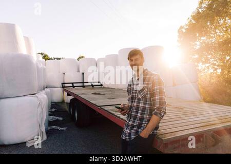 Ritratto di un agricoltore sorridente che si appoggia a un rimorchio con smartphone Foto Stock