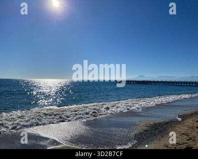 Una luminosa e soleggiata scena costiera che mostra dolci onde che si bagnano su una spiaggia sabbiosa di ciottoli. Un lungo molo di legno si estende lontano nel mare blu calmo. Foto Stock