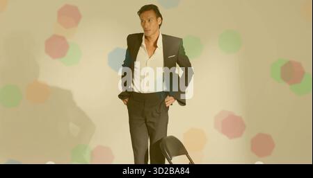 Uomo asiatico in piedi che indossa una tuta dietro una sedia pieghevole in metallo nero in studio con sporgenze esagonali Foto Stock