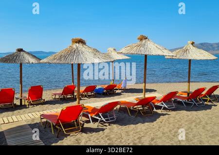 Vista della spiaggia con ombrelloni di paglia e lettini rossi per una vacanza rilassante nella giornata di sole a Skala Kallonis, Lesbo, Grecia Foto Stock