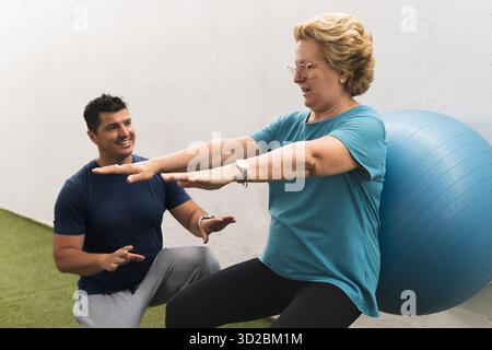 Paziente anziano che fa il fitball squat contro la parete con l'aiuto di un personal trainer. Foto Stock