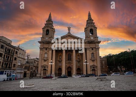 Una chiesa barocca con due campanili si erge in una piazza della Valletta al tramonto, facciata illuminata da lampadine calde, orologi sulle torri, statua sul frontone, automobili A. Foto Stock