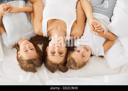 Amate madre e figli, sdraiati a testa in giù sul letto bianco, tenendosi le mani vicine, sorridendo gioiosamente Foto Stock