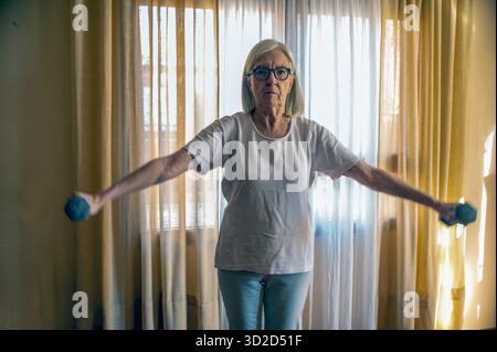 Una donna anziana sta in una stanza soleggiata, le braccia si estendono con i manubri, a metà esercizio. Indossando occhiali e una t-shirt leggera, mostra concentrazione, forza e da Foto Stock