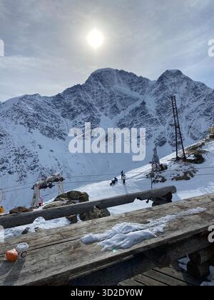 Un soleggiato paesaggio montano di Cheget con una maestosa vetta che domina lo sfondo. In primo piano, una tavola rustica in legno è ricoperta di neve. Foto Stock