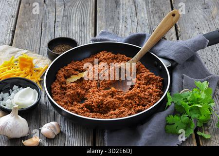 ragù di manzo macinato brasato lentamente in una padella con cucchiaio di legno su un tavolo rustico di legno con formaggio grattugiato, aglio e prezzemolo, vista orizzontale Foto Stock