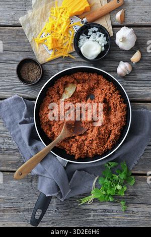 ragù di manzo macinato cotto a fuoco lento in una padella con cucchiaio di legno su un tavolo in legno rustico con formaggio grattugiato, aglio e prezzemolo, vista verticale da a. Foto Stock