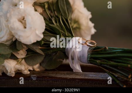 Primo piano di un romantico bouquet floreale bianco con fedi nuziali dorate poggiate su una superficie rustica in legno, simboleggiano l'amore e il matrimonio nella morbida natura Foto Stock