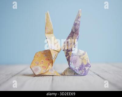 Due colorati coniglietti origami sono rivolti l'uno di fronte all'altro, toccando nasi, su una superficie di legno con sfondo azzurro Foto Stock