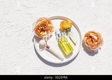 Vista dall'alto del vassoio in ceramica con flacone cosmetico di siero o olio per massaggi e cura della pelle del viso e del corpo, massaggiatore di pietre, rose delicate Foto Stock