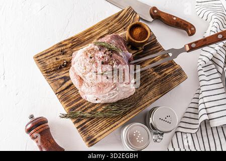 Un grosso pezzo di prosciutto di maiale si trova su un tagliere di carne di legno testurizzato con ingredienti per la cottura. coltello, forchetta per carne, spezie Foto Stock