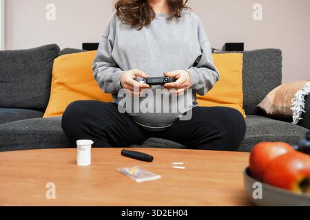 Donna incinta siede sul divano, tenendo un glucometro e preparandosi a controllare il suo livello di zucchero nel sangue per il controllo del diabete gestazionale, CA di salute Foto Stock