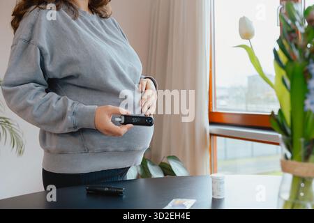 Una donna incinta sta accanto alla finestra, tenendo un glucometro e toccandole la pancia mentre monitora la glicemia per la cura del diabete gestazionale Foto Stock