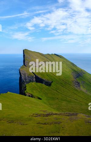 Il bordo della scogliera di Mylingur sorge nettamente dal vibrante paesaggio verde delle Isole Faroe, affacciato sul vasto oceano blu sotto un cielo luminoso. La bellezza della natura è a schermo intero. Foto Stock