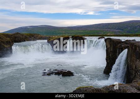 Le cascate di Godafoss crollano drammaticamente sulle scogliere rocciose, circondate da lussureggianti colline verdi sotto un vasto cielo in Islanda. Il potente flusso d'acqua mostra la natura cruda bellezza e tranquillità Foto Stock