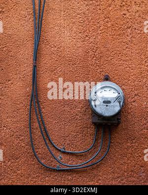 Il misuratore elettrico montato su una parete in cemento color terracotta mostra semplici componenti industriali contro superfici ruvide, adatto per l'energia, u Foto Stock