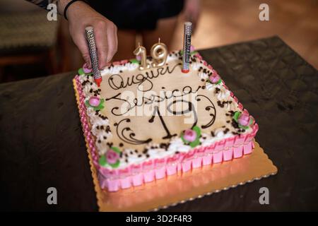 Metti a mano una candela scintillante in una torta di compleanno rosa e bianca decorata per la celebrazione del 19° compleanno di jessica, con shav al cioccolato Foto Stock