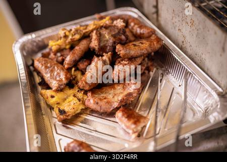 Salsicce cotte, pollo e altre carni riposate in un vassoio di alluminio usa e getta con segni di griglia, pronto per essere servito a una festa barbecue all'aperto Foto Stock